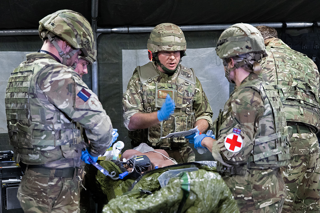 TMW medics in camouflage uniforms, body armour, and combat helmets treat a medical simulation dummy inside a tent. The medic in the centre wears blue gloves and directs the team while holding paperwork, while a female medic on the right features a prominent red cross patch on her shoulder arm.